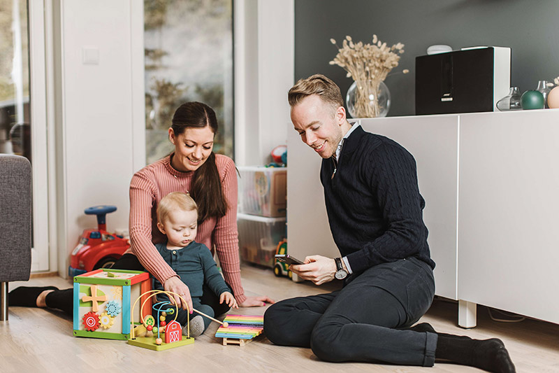 To foreldre og en baby leker på gulvet hjemme mens mannen sjekker Fjordkraft-appen